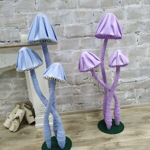 May include: Two sets of whimsical mushroom sculptures. One set is light blue, the other is purple. Each mushroom has a cap and stem, with the stems intertwined. The mushrooms are on green circular bases, set against a white brick wall.