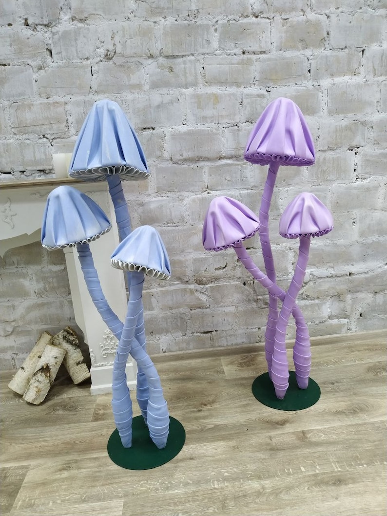 May include: Two sets of whimsical mushroom sculptures. One set is light blue, the other is purple. Each mushroom has a cap and stem, with the stems intertwined. The mushrooms are on green circular bases, set against a white brick wall.