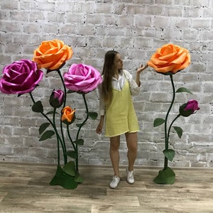 May include: Large, colorful paper roses in shades of orange, pink, and magenta. The oversized flowers are mounted on green stems with leaves. The arrangement is set against a white brick wall and a wooden floor.