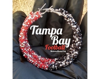 May include: A pair of hoop earrings, half red and white, half black and silver, with the text "Tampa Bay Football". The earrings are covered in small, sparkling crystals.