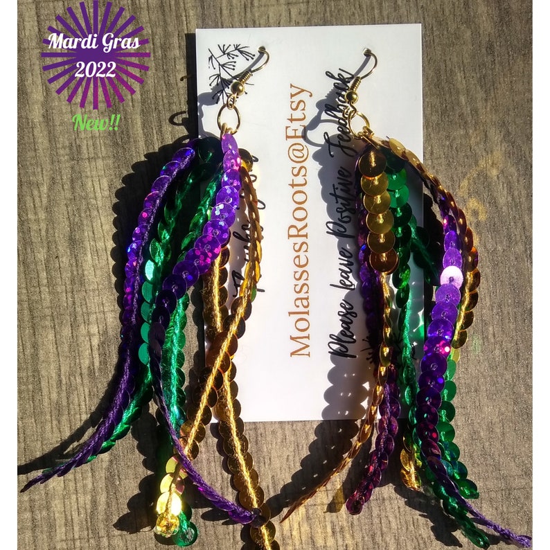 May include: Two pairs of Mardi Gras themed dangle earrings. Each earring is made of gold, green, and purple sequins. The earrings are hanging from a card that says 'MolassesRoots@Etsy' and 'Please leave positive feedback'. The card also has a small drawing of a plant on it.