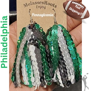 May include: Green, white, and black sequined tassel earrings. The earrings are in the shape of a tassel and are made of sequins. The earrings are hanging from a silver hook.