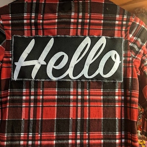 May include: Red and black plaid flannel shirt with a black patch sewn on the back that says "Hello" in white lettering.