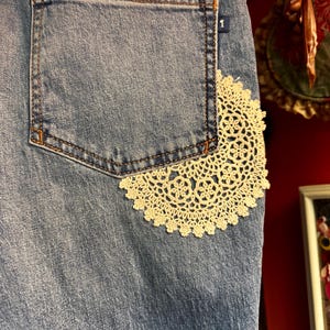 May include: Close-up of a light blue denim garment with a back pocket. A cream-colored, circular lace doily is sewn onto the denim, partially covering the pocket. The pocket has orange stitching.