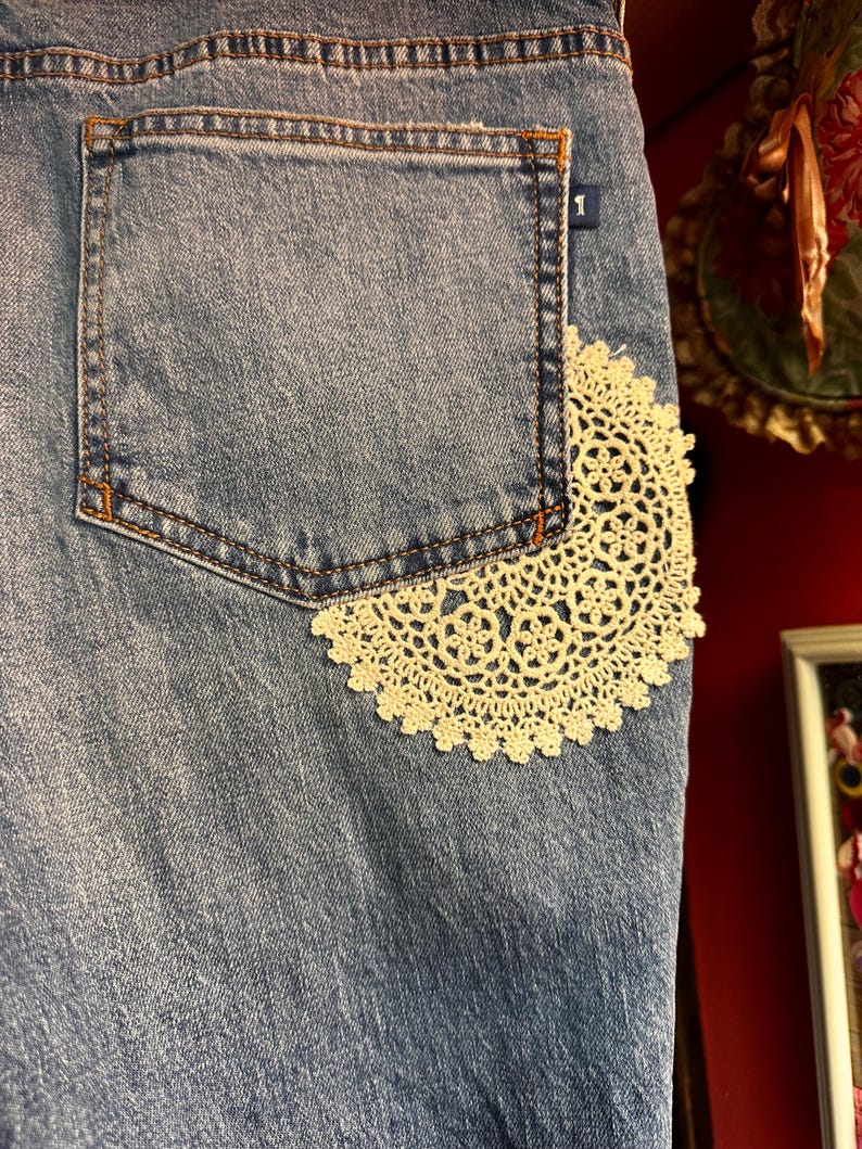 May include: Close-up of a light blue denim garment with a back pocket. A cream-colored, circular lace doily is sewn onto the denim, partially covering the pocket. The pocket has orange stitching.