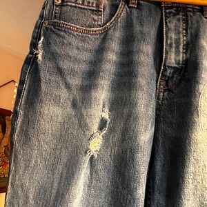 May include: Close-up of blue denim jeans with distressed details. The jeans have a classic five-pocket design, a button and zipper closure, and a light wash. The jeans are hanging on a wooden hanger with "Macy's California" printed on it.