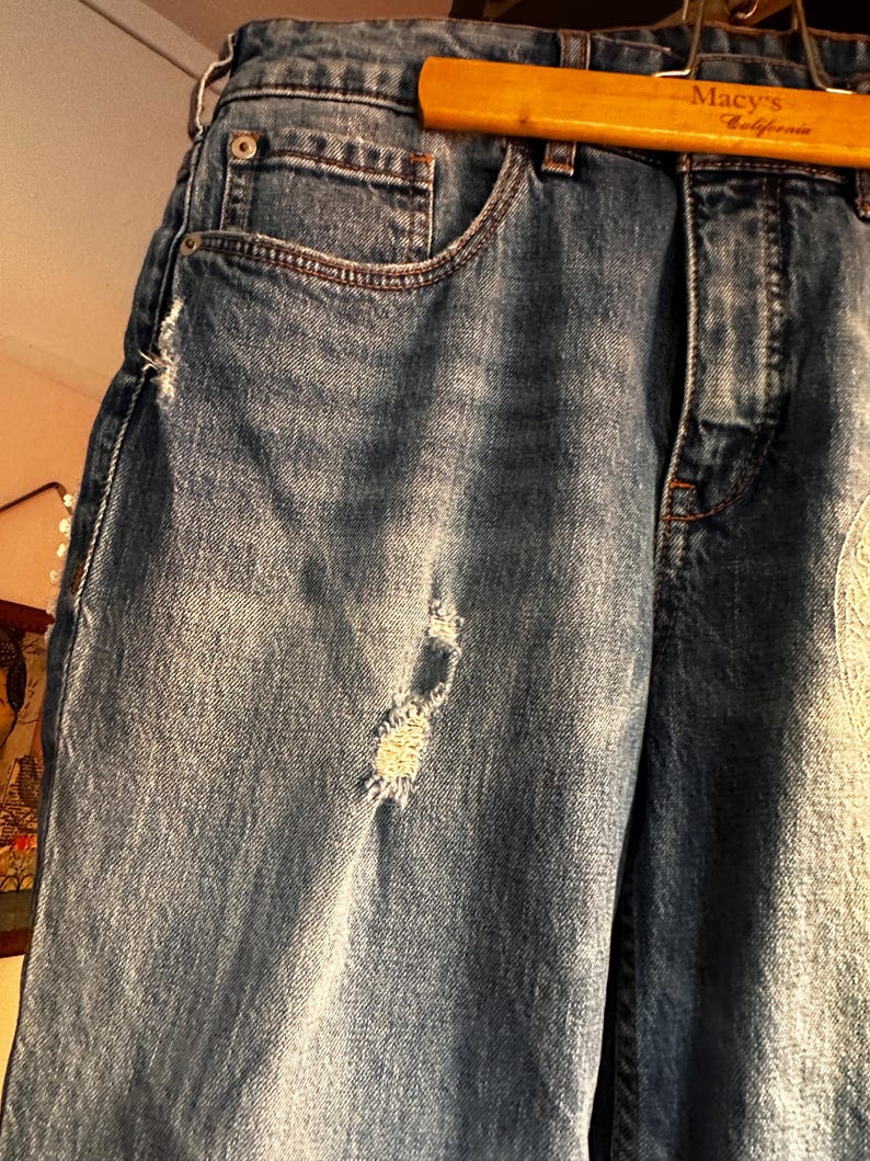 May include: Close-up of blue denim jeans with distressed details. The jeans have a classic five-pocket design, a button and zipper closure, and a light wash. The jeans are hanging on a wooden hanger with "Macy's California" printed on it.