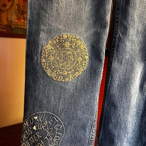 May include: Close-up of blue denim jeans with decorative patches. One patch is a gold-colored, circular lace design. Another patch features white embroidered text including names like Cleopatra and Romeo.