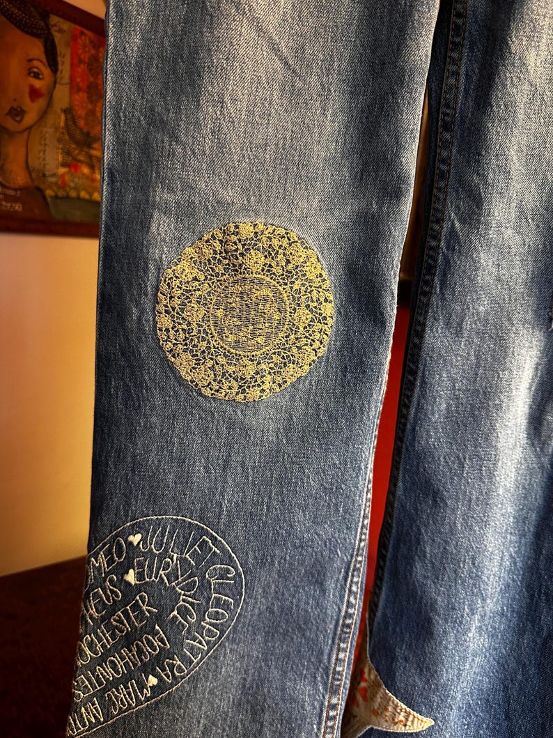 May include: Close-up of blue denim jeans with decorative patches. One patch is a gold-colored, circular lace design. Another patch features white embroidered text including names like Cleopatra and Romeo.