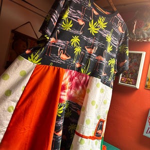 May include: A patchwork dress featuring a black top with a flamingo and palm tree print. The skirt has panels of orange, tie-dye, and white fabric with green polka dots. A unique, colorful, and artistic garment.