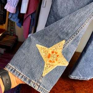 May include: Blue denim jeans with a decorative star patch on the lower leg. The star is beige with a floral and heart pattern in orange and red. The jeans are held by a person.