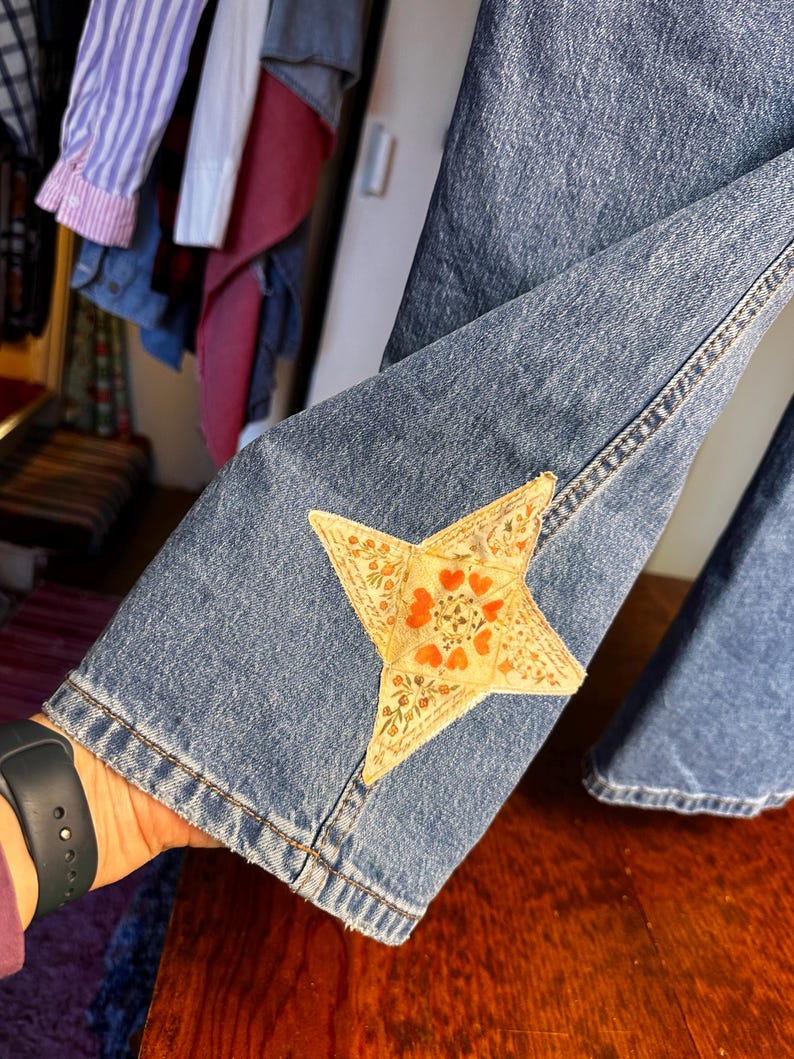 May include: Blue denim jeans with a decorative star patch on the lower leg. The star is beige with a floral and heart pattern in orange and red. The jeans are held by a person.