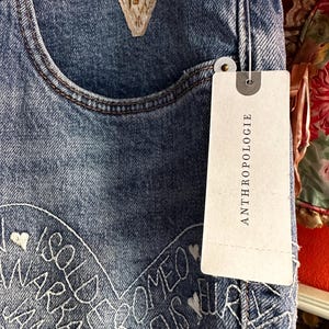 May include: Close-up of blue denim jeans with white embroidered text and heart details. The jeans have a decorative floral patch near the pocket and a tag that reads "ANTHROPOLOGIE".