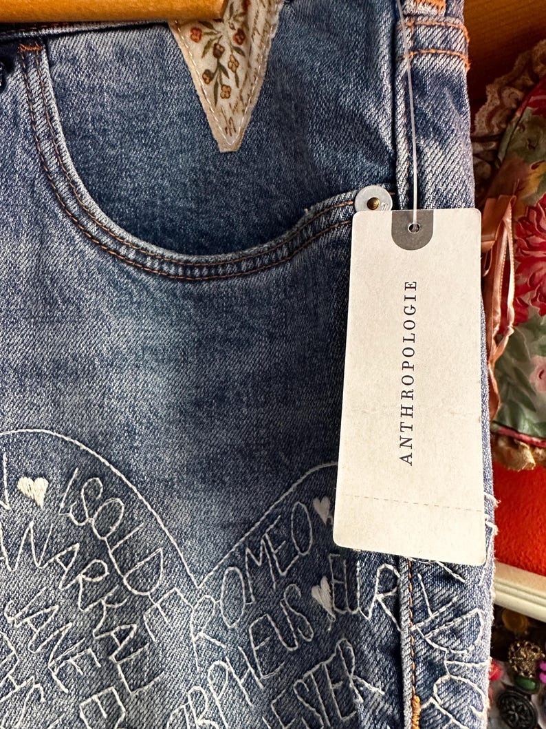 May include: Close-up of blue denim jeans with white embroidered text and heart details. The jeans have a decorative floral patch near the pocket and a tag that reads "ANTHROPOLOGIE".