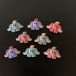 May include: A collection of small, colorful Dumbo-themed embellishments. Each elephant figure has large ears and wears a hat. The elephants are in shades of blue, pink, and purple, with hats in yellow, pink, and red.
