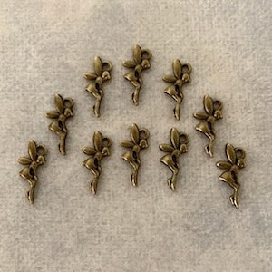 May include: A collection of ten small, antique gold-toned metal charms shaped like fairies with wings. The charms are arranged in a scattered pattern on a light beige surface.