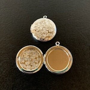 May include: Two silver-colored round lockets with floral patterns. One locket is open, revealing a clear interior with a floral design. The other locket is closed. Both have a small loop at the top for a chain.
