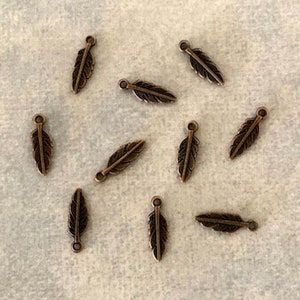 May include: A collection of 12 bronze-colored metal feather charms. The charms are small and delicate, with a simple design. They are perfect for adding a touch of whimsy to jewelry or crafts.