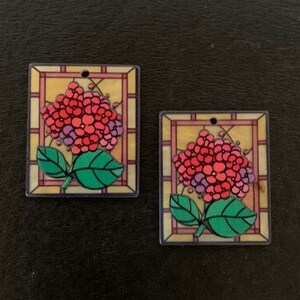 May include: Two square-shaped stained glass style pendants with a pink and purple flower design. The flower is surrounded by a green leaf and a yellow background.