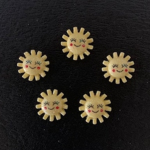 May include: Five small, yellow sun-shaped embellishments with smiling faces and red cheeks are arranged on a black surface. Each sun has a detailed, textured design with rays extending outward. These decorative elements could be used for crafts or embellishments.