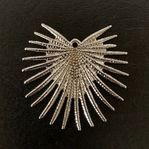 May include: Silver heart-shaped pendant with a starburst design. The pendant has a hole at the top for attaching a chain or cord.