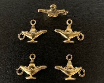 May include: Five gold-colored genie lamp charms. Each charm features a curved spout, a handle, and a small loop at the top for attaching to jewelry or other crafts. The lamps are arranged on a dark surface.