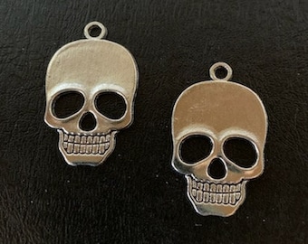 May include: Two silver-colored skull charms with large eye sockets and detailed teeth. Each charm has a small loop at the top for attaching to jewelry or other crafts. The skulls are displayed against a dark background.