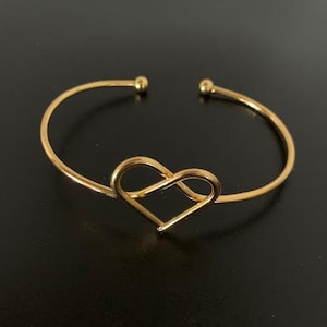 May include: A gold-colored open cuff bracelet with a heart-shaped centerpiece. The bracelet has a simple, elegant design, with a smooth, polished finish. The heart is formed by a continuous line, creating an open, airy feel. The bracelet is likely made of metal.