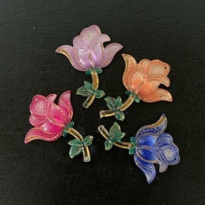 May include: Five plastic flower embellishments in various colors, including pink, purple, orange, and blue. The flowers have a 3D design and are perfect for adding a touch of whimsy to crafts and projects.