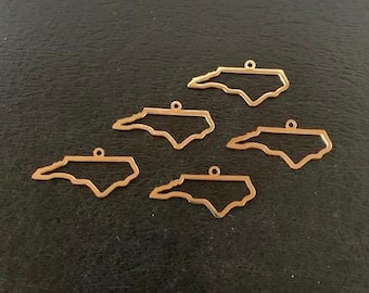 May include: Six gold-coloured outline charms in the shape of North Carolina. Each charm has a small loop at the top for attaching to a chain or other jewellery component. The charms are arranged on a dark surface.