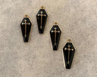 May include: Four black coffin-shaped charms with gold cross accents. The charms are on a light beige background.