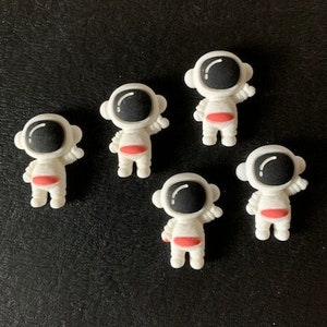 May include: Five white astronaut figurines with black helmets and red accents. The figurines are standing on a black surface.
