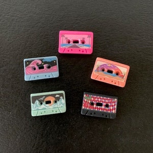 May include: Five colorful cassette tape buttons with different designs. The buttons are pink, blue, green, black, and red. The designs include palm trees, waves, and a checkered pattern.