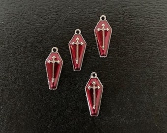 May include: Four red coffin-shaped charms with silver-colored trim and a cross design. Each charm has a small loop at the top for attaching to jewelry or other crafts. The charms are displayed on a dark, textured surface.