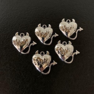 May include: Five silver charm pendants shaped like hearts with a devil tail. Each heart has a flame design and the word "evil" written inside.