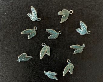 May include: A collection of ten small, antique-looking metal leaf charms. The leaves are a light blue-green color and have a slightly distressed finish. Each leaf charm has a small loop at the top for attaching to a necklace, bracelet, or other jewelry.