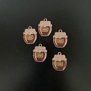 May include: Five gold-colored metal charms shaped like honey jars with pink lids. The jars are filled with a dark red substance and have a gold label that says "Honey".