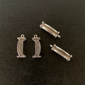 May include: Three silver metal charms, two are shaped like a building with a detailed design and one is a rectangular shape with a clasp.