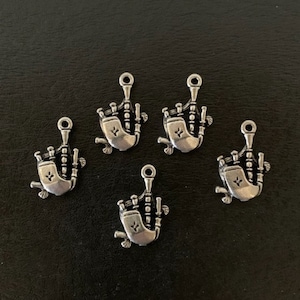 May include: Five silver-toned bagpipe charms. Each charm features a detailed miniature bagpipe design with a loop at the top for attaching to jewelry or other crafts. The charms are displayed on a dark surface.