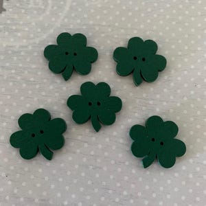 May include: Five dark green wooden shamrock buttons. Each button has a three-leaf clover shape with two small holes. The buttons are arranged on a white surface with small, evenly spaced gray dots.