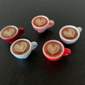 May include: Five miniature coffee cups with latte art in the shape of a heart. The cups are in various colors, including red, pink, blue, and white. The coffee has a brown color, and the heart design is in a lighter shade.