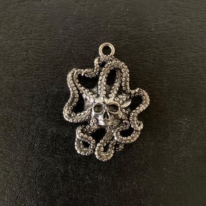 May include: A silver-colored pendant featuring a skull at the center, surrounded by octopus tentacles. The pendant has a loop at the top for attaching to a chain. The design is detailed and has a gothic or nautical theme.