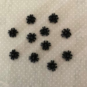 May include: Twelve black spider-shaped embellishments on a white background with a polka dot pattern.