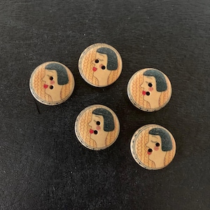 May include: Five round wooden buttons with a vintage design. Each button features a profile of a woman with a red lip, black hair, and a cream-colored face. The background has a wavy pattern.