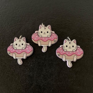 May include: Three cat-shaped charms. Each charm features a white cat floating in a pink donut with white star sprinkles. The donuts have a light brown base. Each charm has a silver-colored loop at the top.