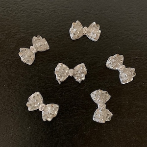 May include: Six silver glitter bow embellishments. Each bow is made of small, sparkly silver pieces and has a clear rhinestone in the center.