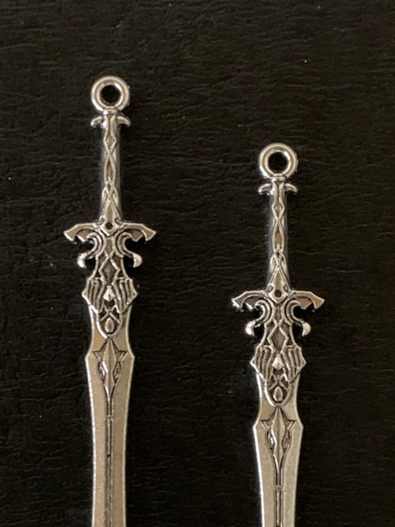 Pu&ograve; includere: Due ciondoli a forma di spada color argento con disegni intricati. Ogni ciondolo presenta un'elsa e una lama dettagliate, con un anello nella parte superiore per il fissaggio a gioielli o altri lavori artigianali. Le spade sono su sfondo scuro.
