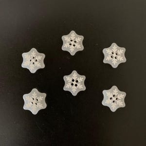 May include: Six light blue, snowflake-shaped buttons arranged in a circular pattern. Each button features a white, intricate, six-pointed snowflake design embossed on the surface with four small holes in the center for sewing attachment.