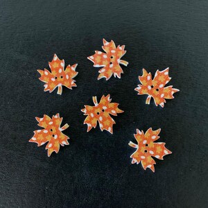 May include: Six wooden leaf-shaped buttons. Each button is orange with a white and orange plaid pattern and two small holes for sewing. The buttons are arranged on a black surface.