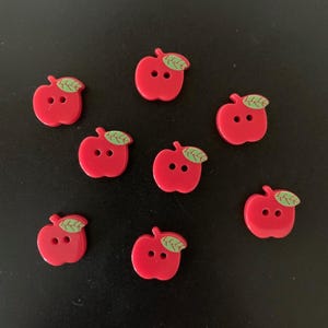 May include: A collection of seven red apple-shaped buttons, each featuring two central holes for attachment and a small green leaf detail at the top. The buttons are arranged in a scattered pattern, showcasing their glossy finish and uniform design.